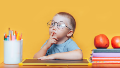 A young child wearing glasses sits at a desk with a finger on their lips, surrounded by colored pencils, books, and apples, against a bright yellow background.