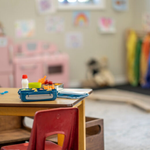 Classroom with a focus on a desk with lunch items in the foreground, colorful jackets and artwork visible in the blurred background.