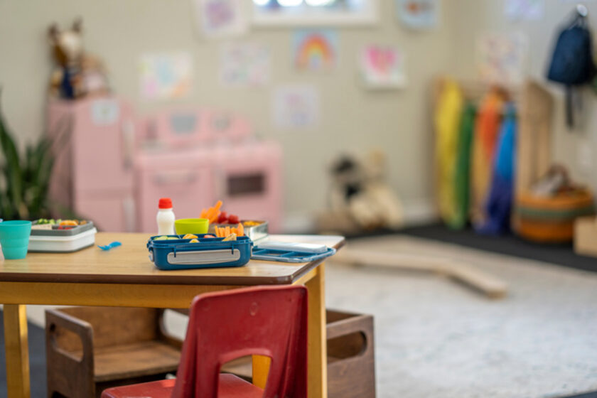 Classroom with a focus on a desk with lunch items in the foreground, colorful jackets and artwork visible in the blurred background.