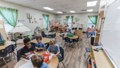 Children sit and work at tables in a classroom decorated with plants and wall art, while others play or read in different activity areas around the room.